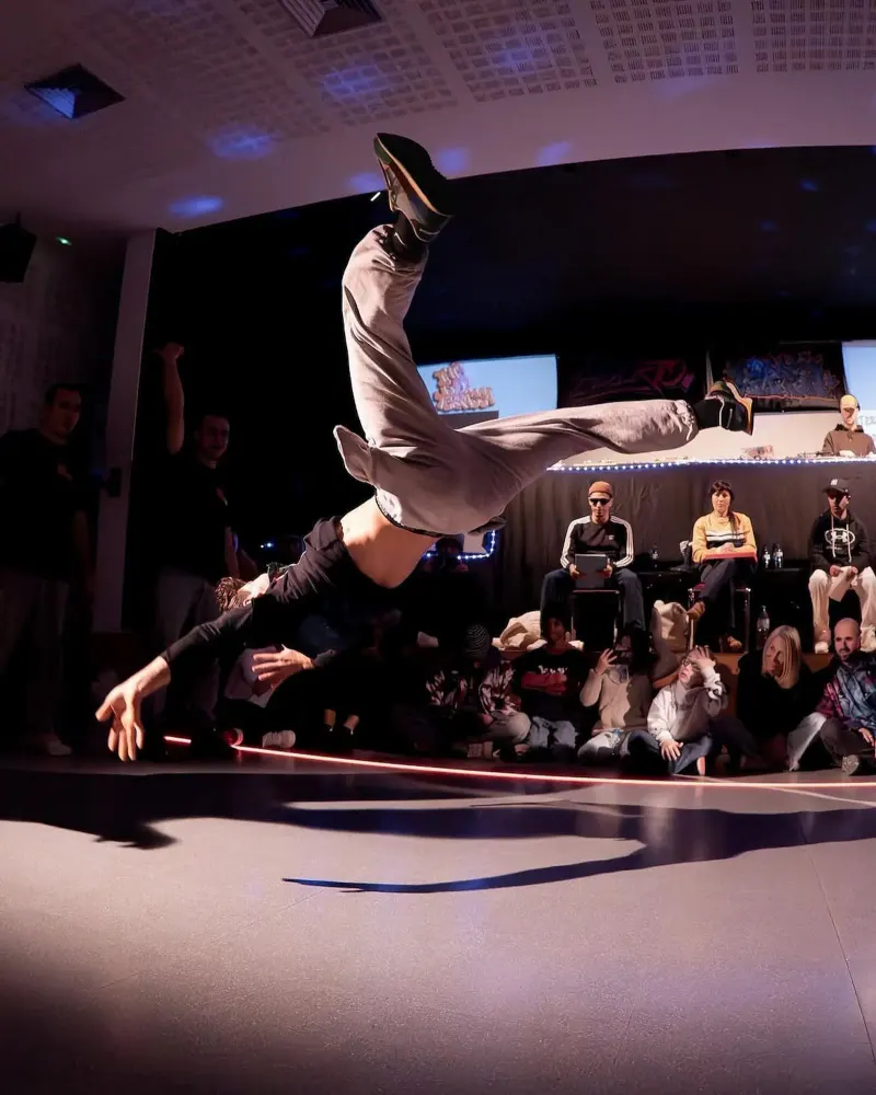 El bboy Twista fent un moviment aeri en una batalla, amb el públic assegut a terra