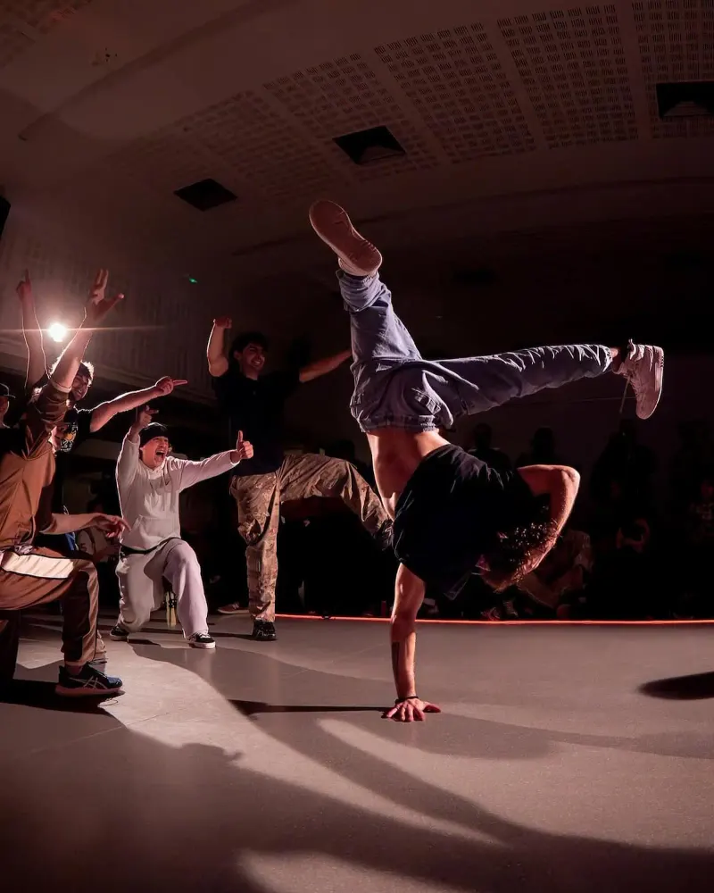 Un bboy recolzat en una mà amb les cames enlaire mentre el seu crew l'anima
